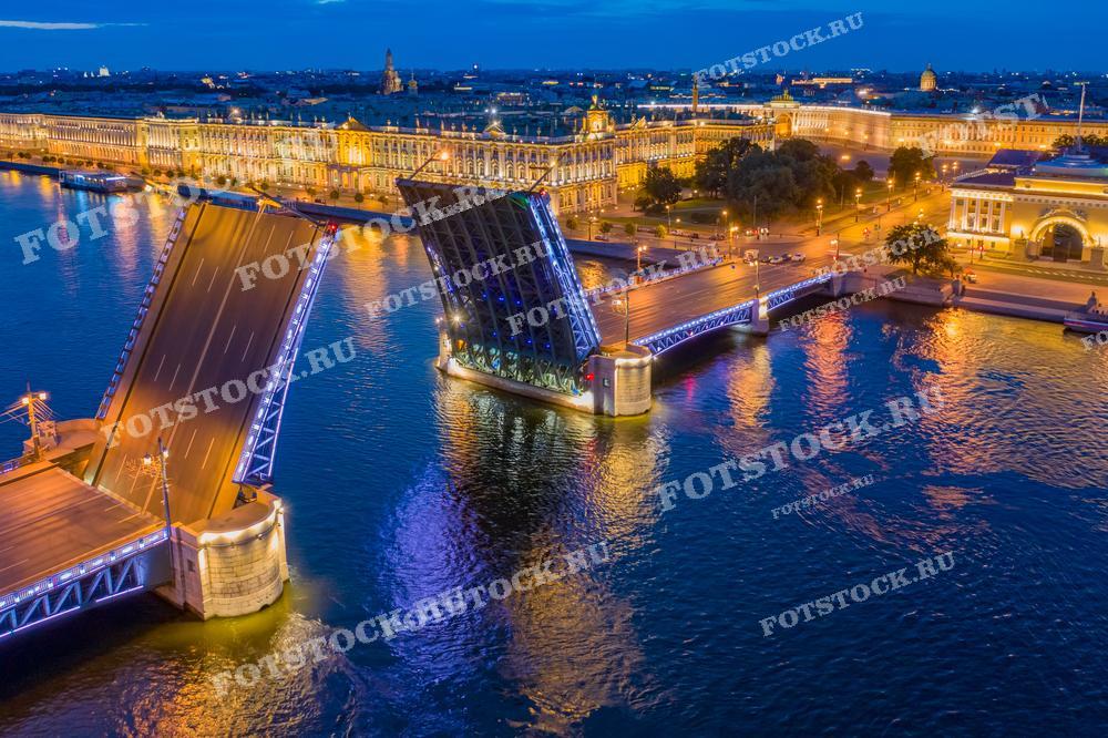 На фотографии изображен Дворцовый мост в Санкт-Петербурге, разведенный ночью. На заднем плане ярко освещен Эрмитаж, один из самых известных музеев мира. Река Нева отражает огни мостов и зданий, создавая красивую ночную панораму города.